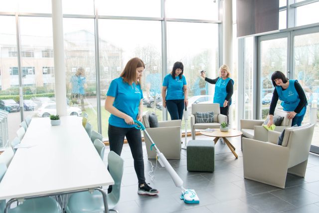 Four Archway Facilities cleaning operatives in blue uniforms performing deep cleaning in a bright, modern UK office lounge. One uses a high-efficiency floor steam cleaner, another vacuums, and others wipe down soft seating and surfaces to improve indoor air quality.