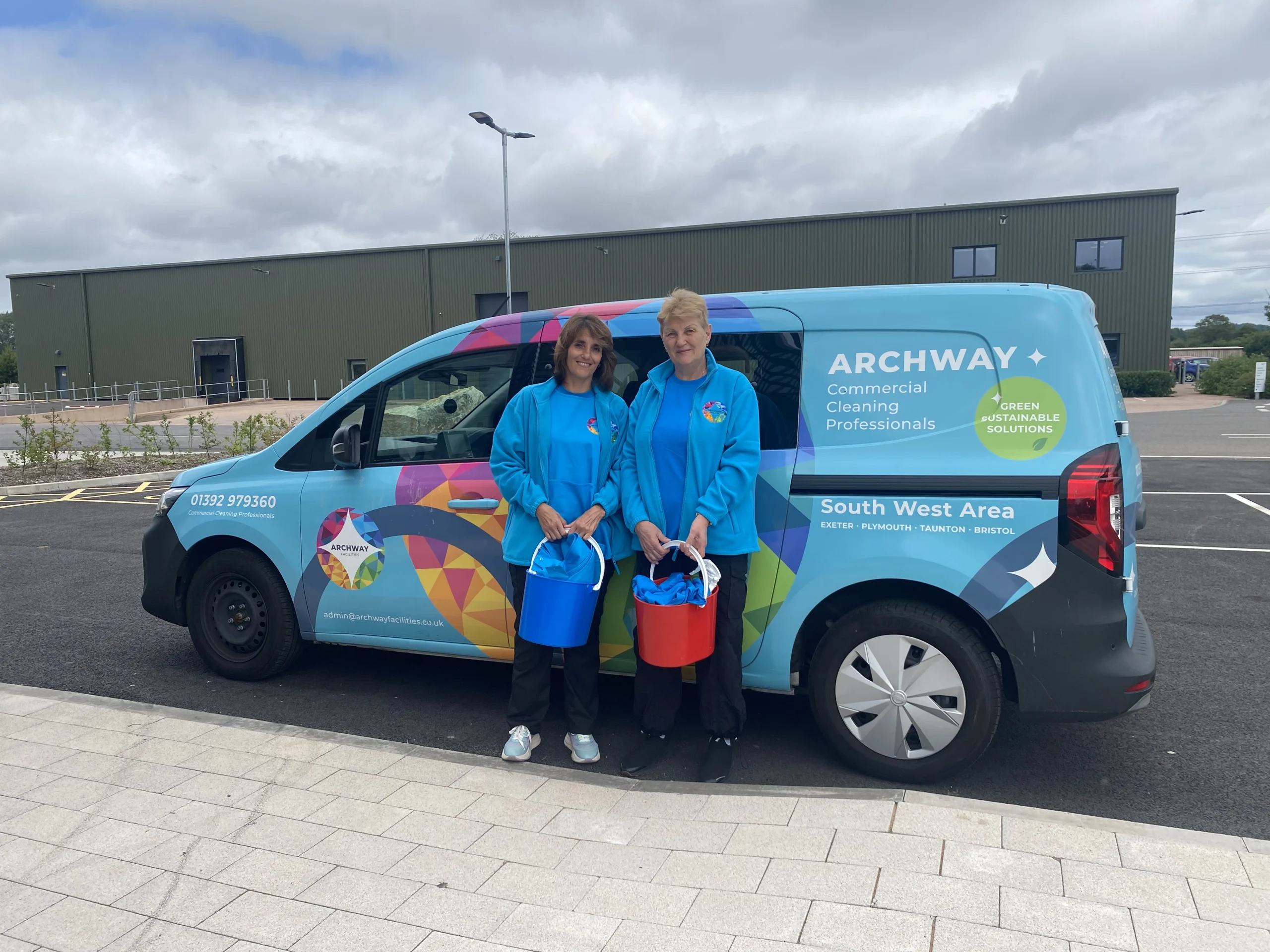 Two Archway Facilities team members in blue uniforms holding cleaning supplies beside a branded company vehicle outside a commercial building.